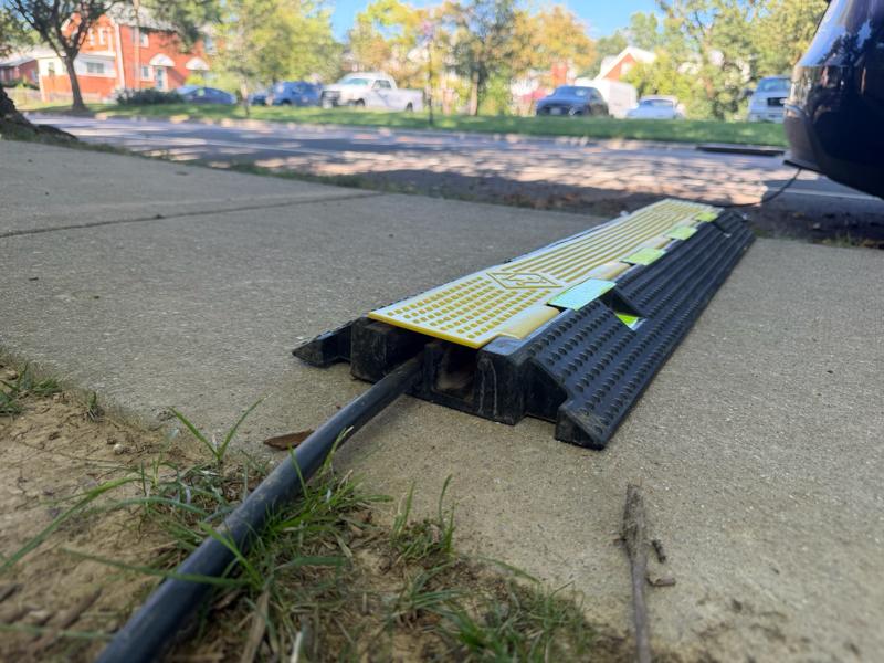 Low-angle close-up of the cable protector on the sidewalk: the charging cable enters an enclosed channel under a hinged yellow lid, with beveled, dimpled edges on both sides and a parked car at the curb behind it.