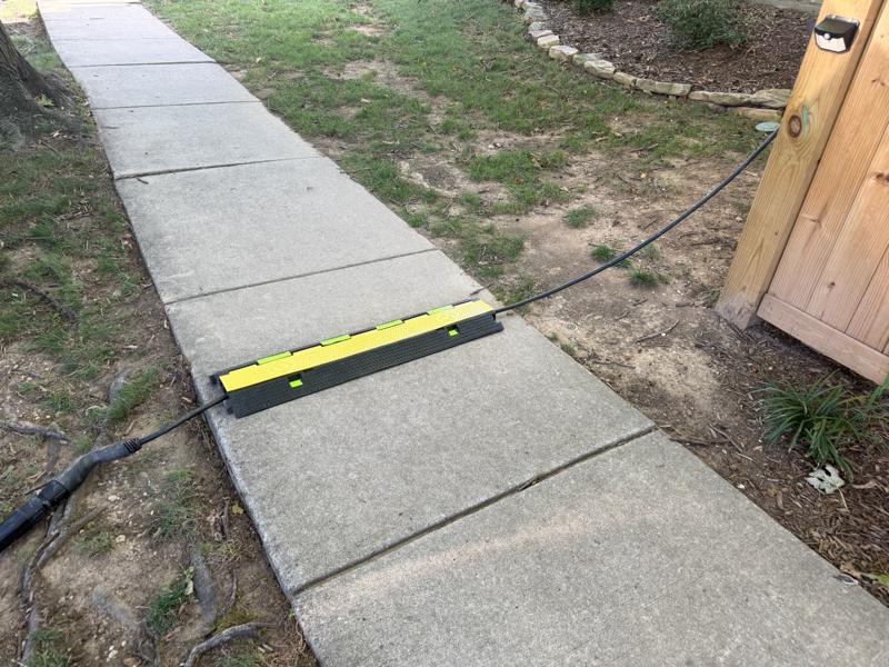 A yellow-and-black pedestrian cable protector lying flat across a concrete sidewalk, enclosing a charging cable that runs from a wooden fence on the house side to the tree lawn at the curb.